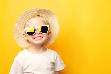 Portrait of a cute blond baby boy with a happy face smile in a straw hat and sunglasses on a yellow background looking at the camera. card with place for text
