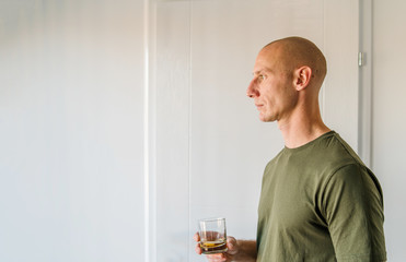 Portrait of young caucasian man good looking with short hair wearing green t shirt holding a glass of brandy or whiskey alcohol drink standing in front of white wall at home