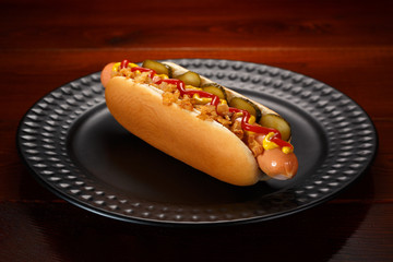 Danish hot dog with pickled cucumbers, fried onions, ketchup and mustard. On a black plate.