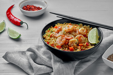 Shrimp fried rice with vegetables, green onions and lime in a black pottery bowl on a tablecloth. Traditional Thai meal with sesame on a white table.