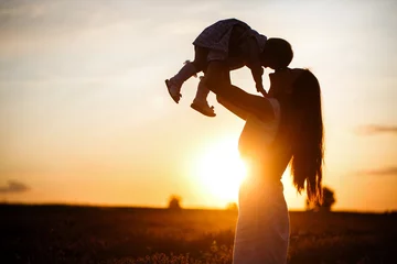 Fotobehang A mother lifts a toddler child in the air above a picturesque sunset sky. A woman and a little girl in a field of lavender flowers. Copy space © Yulia Sugarbox