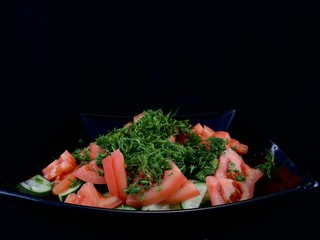 Vegetable salad of cucumbers, tomatoes and herbs on a black background. Finely chopped vegetables in a salad bowl, close-up. Concept: healthy diet, fresh vegetables, vitamins.