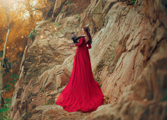 Luxury sexy mysterious power brunette woman in red dress long train. black raven on hand. Fabolous photography. Creative outfit design. Background autumn nature rocks mountains. Rear view looking back