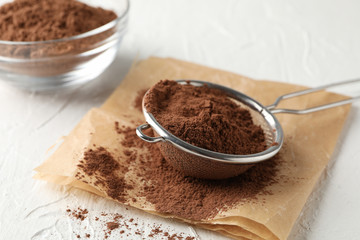 Glass bowl and strainer with cocoa powder, baking paper on white background, close up