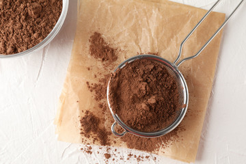 Glass bowl and strainer with cocoa powder, baking paper on white background, close up