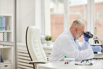 Mature medical specialist with grey hair sitting at the table and analyzing sample through the microscope at office