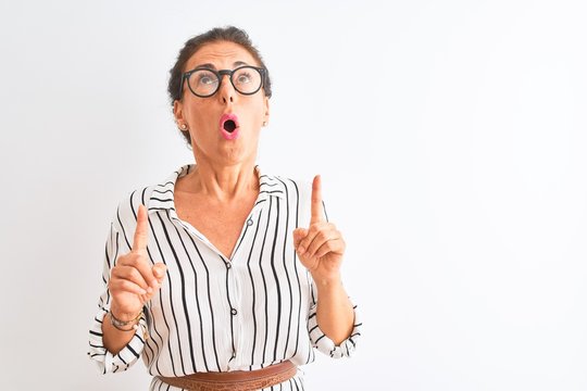 Middle age businesswoman wearing striped dress and glasses over isolated white background amazed and surprised looking up and pointing with fingers and raised arms.
