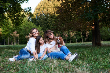 Two young blond women and one brunette girl, wearing jeans and white t-shirts, sitting on green grass in park, playing with cavalier king charles spaniel, smiling, laughing. Leisure time in summer.