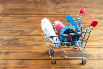 Tailor needles, threads, seam ripper and measuring tape in a shopping cart with wooden background. Cost sewing supplies.