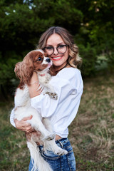 Young pretty blond woman with curly hair, wearing eyeglasses and white shirt, holding small white and brown dog in park in summer, smiling. Cavalier king charles spaniel with his owner on a walk.