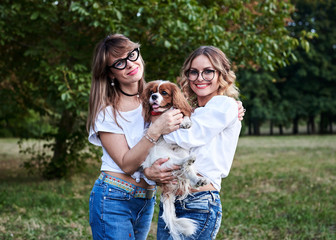 Two young blond women, wearing white t-shirts,, holding small white and brown dog in park in summer, smiling. Cavalier king charles spaniel with his owners on a walk.