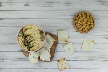 hummus is a traditional dish of eastern cuisine in a pink plate, on a white background and wooden board . homemade of chickpea, sesame paste, lemon, decorated with seeds and crisps. flat lay.