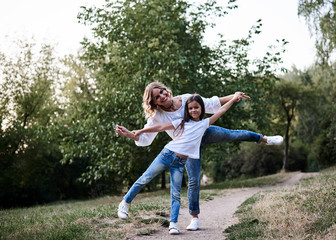 Naklejka na ściany i meble Young blond mother posing for picture with her brunette daughter in the park in summer. Woman and girl, wearing blue jeans and white t-shirts, having fun, laughing, smiling. Family leisure fun time.