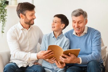 Boy Reading Book Sitting With Father And Grandfather On Sofa