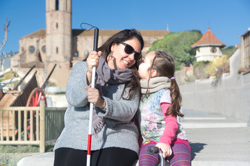 Blind mother with a white cane and her daughter talking sitting on a bench in the park and smiling. Visually impaired lifestyle concept with empty copy space.