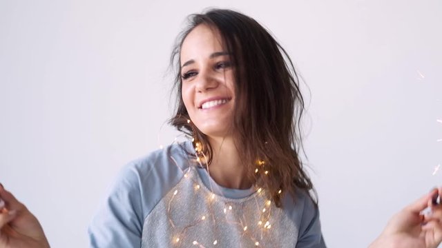 Festive mood with sparklers. Attractive girl in pajamas with sparklers and a garland waiting for the holidays on a white background