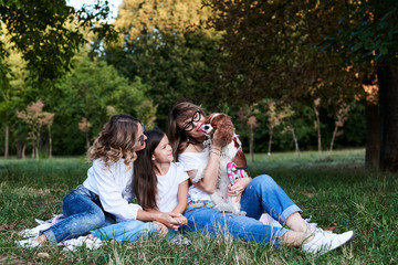 Two young blond women and one brunette girl, wearing jeans and white t-shirts, sitting on green grass in park, playing with cavalier king charles spaniel, smiling, laughing. Leisure time in summer.