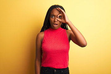 African american woman wearing red casual t-shirt standing over isolated yellow background doing ok gesture shocked with surprised face, eye looking through fingers. Unbelieving expression.