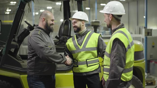 Portrait of bearded Caucasian man getting out the tow tractor and greeting his colleagues in white helmets and green vests. Workers in manufacture plant. Professional, working, loader, employee, mill.