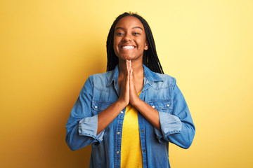 Young african american woman wearing denim shirt standing over isolated yellow background praying...
