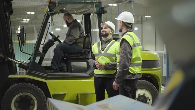 Two Caucasian men talking as standing next to the tow tractor. Bearded driver sitting in cabin at the background. Workers in manufacture plant. Professional, working, loader, employee, mill.