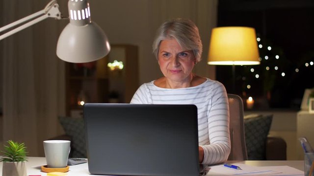technology, old age and people concept - happy senior woman with laptop computer drinking coffee at home in evening