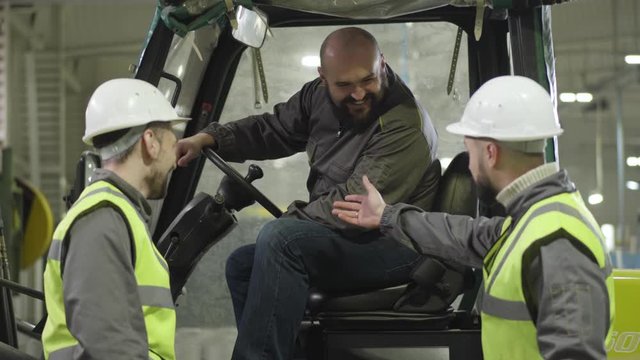Two male Caucasian workers in helmets and vests talking to bearded colleague sitting in tow tractor. Plant employees working indoors. Manufacture, loader, production.