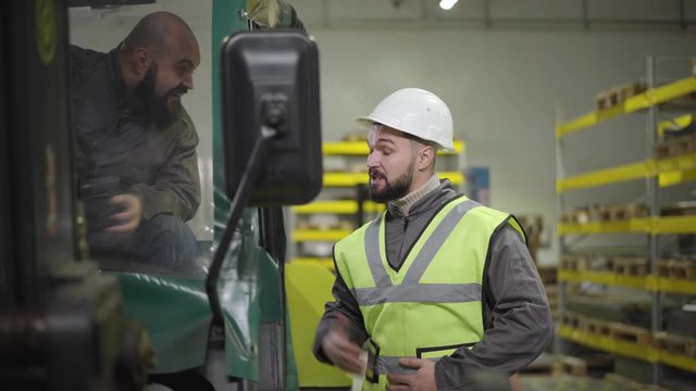 Portrait of tow tractor driver talking to Caucasian man in helmet and vest. Workers having break at production site. Manufacture, working, warehouse, industry.