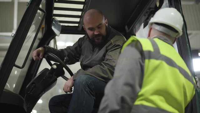 Portrait of bearded bald Caucasian man sitting in tow tractor cabin and talking to worker in helmet and vest. Employees having break at warehouse. Manufacture, working, warehouse, industry.