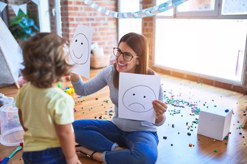 Young beautiful psicologist and toddler doing therapy using emoji emotions at kindergarten