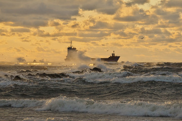 dramatic view to vessel leaving a harbor and passing a pier, breaking through stormy waves, brightly lit by the sunset light 