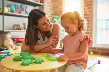 Fototapeta premium Beautiful teacher and blond student toddler girl drawing using digital board at kindergarten