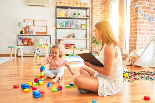 Beautiful psycologist and blond toddler girl doing therapy building tower using plastic blocks at kindergarten