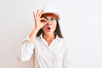 Young beautiful architect woman wearing helmet and glasses over isolated white background doing ok gesture shocked with surprised face, eye looking through fingers. Unbelieving expression.