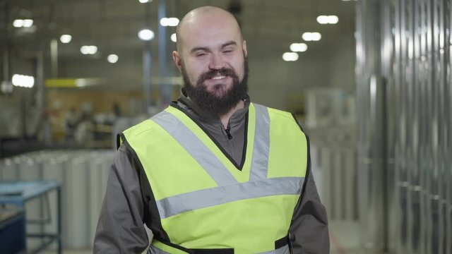 Portrait of bald Caucasian man with beard standing at warehouse, looking at camera and smiling. Happy worker at his workplace at production site. Plant, mill, manufacturing, employee.