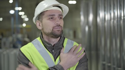 Exhausted Caucasian man in helmet and vest rubbing eyes and stretching at production site. Tired young worker standing at the background of steel metal pipes. Manufacture, industry, steel plant.