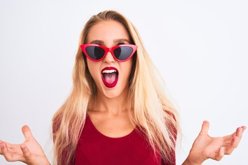 Young beautiful woman wearing red t-shirt and sunglasses over isolated white background crazy and mad shouting and yelling with aggressive expression and arms raised. Frustration concept.