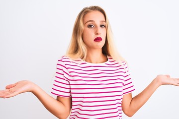 Young beautiful woman wearing pink striped t-shirt standing over isolated white background clueless and confused expression with arms and hands raised. Doubt concept.