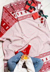 Fototapeta premium Female in white woolen sweater holding in hands a cup of coffee on plaid. On legs red Christmas knitted socks. Preparing gifts. Unboxing day. Background layout for holidays mood copy space.