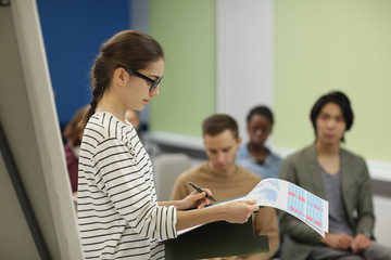 College student in eyeglasses standing and reading her report to other students during seminar