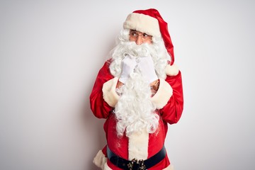 Middle age handsome man wearing Santa costume standing over isolated white background laughing nervous and excited with hands on chin looking to the side