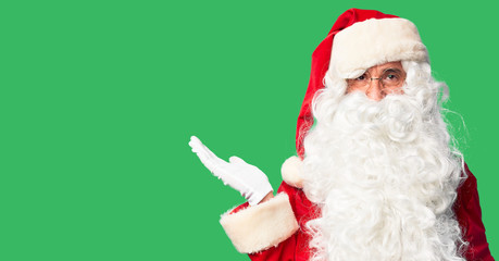 Middle age handsome man wearing Santa Claus costume and beard standing smiling cheerful presenting and pointing with palm of hand looking at the camera.