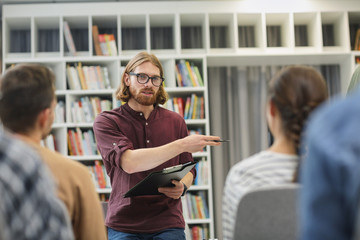 Young teacher holding report and explaining the new material to students during a lesson in the library