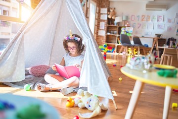 Beautiful toddler wearing glasses and unicorn diadem sitting on the floor inside tipi reading book...