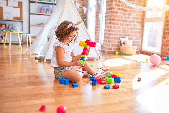 Beautiful toddler wearing glasses and unicorn diadem sitting playing with building blocks at kindergarten