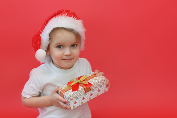 Portrait of smiling joyful child boy in Santa red hat holding Christmas gift in hand over the red background