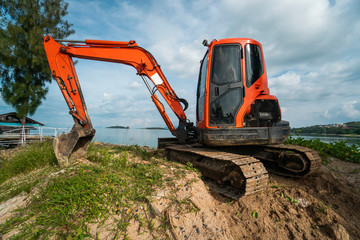 Small orange excavator on a ground against blue sky and sea for a works on construction site. Small tracked excavator standing on a ground with a blue sea on background. Heavy industry.