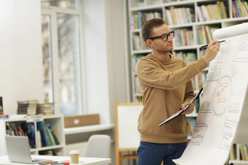 Young man in eyeglasses drawing graphics on whiteboard during business presentation in the library