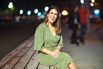 Fototapeta premium Young beautiful girl smiling happy and confident relaxing on bench at the promenade, sitting with a smile on face around night lights bokeh