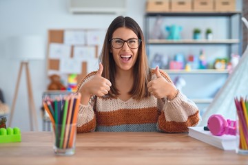 Young beautiful teacher woman wearing sweater and glasses sitting on desk at kindergarten success...